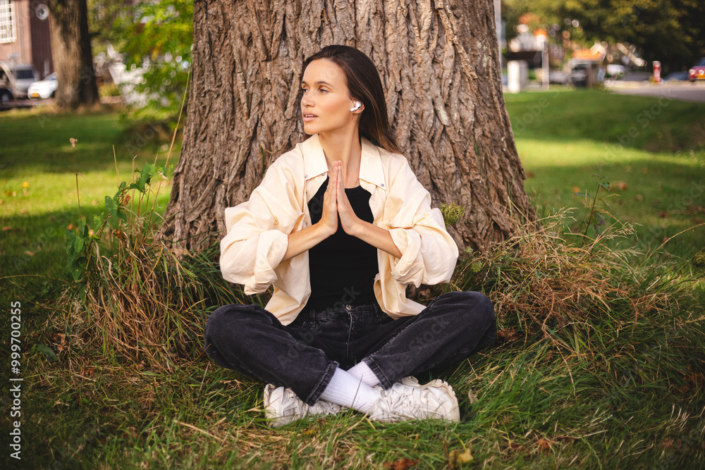 Thankful woman sitting in park, her hands in namaste gesture. Smiling peaceful young woman practicing yoga and meditation, listen mantra, music. Woman in earbuds sitting under trees, meditating. Thankful woman sitting in park, her hands in namaste gesture. Smiling peaceful young woman practicing yoga and meditation, listen mantra, music. Woman in earbuds sitting under trees, meditating.