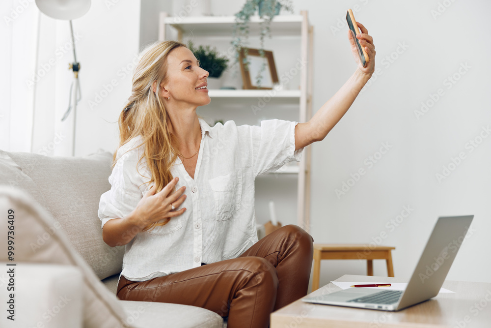 Woman taking a selfie on a couch with her phone while working on a laptop in a cozy living room Woman taking a selfie on a couch with her phone while working on a laptop in a cozy living room