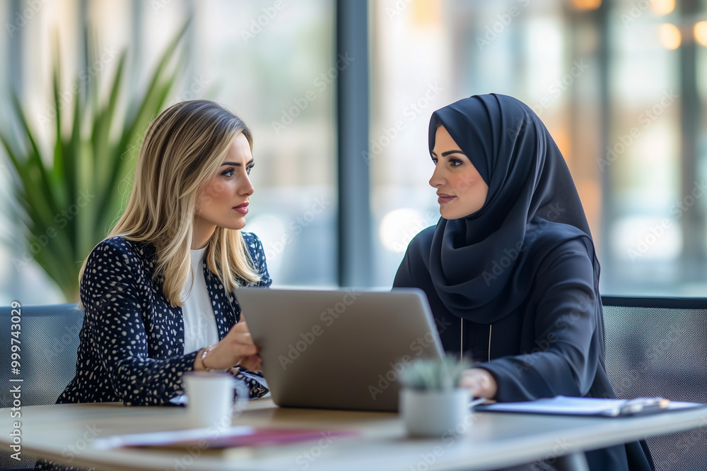 Concentrated young arabian woman in hijab sitting with smiling colleague at table, looking at computer screen, explaining new company software. Focused team leader training millennial female intern. Concentrated young arabian woman in hijab sitting with smiling colleague at table, looking at computer screen, explaining new company software. Focused team leader training millennial female intern.