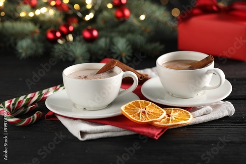 Tasty hot cocoa drink with cinnamon sticks in cups, dry orange and candy canes on black wooden table