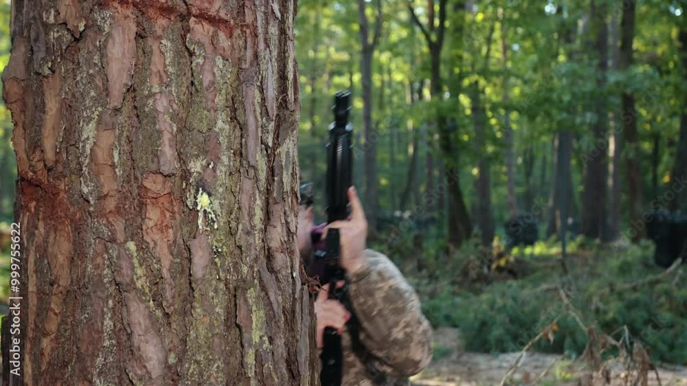 Dense woodland, outdoor game, combat attire. Man wearing camouflage clothing and marker on his head is positioned against tree, carefully targeting Laser tag gun and hitting enemy in forested area Dense woodland, outdoor game, combat attire. Man wearing camouflage clothing and marker on his head is positioned against tree, carefully targeting Laser tag gun and hitting enemy in forested area