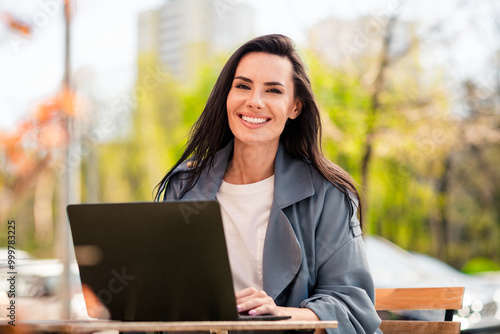 Photo of cheerful pretty lady dressed grey jacket smiling sitting cafe communicating modern device outdoors town street