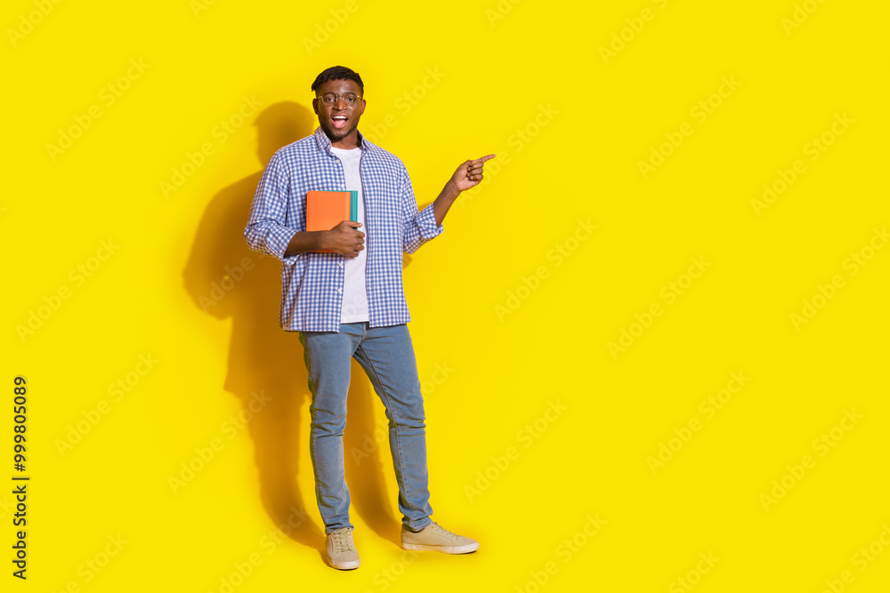 Fototapeta premium Full length photo of funky cool guy dressed checkered shirt holding book pointing emtpy space isolated yellow color background