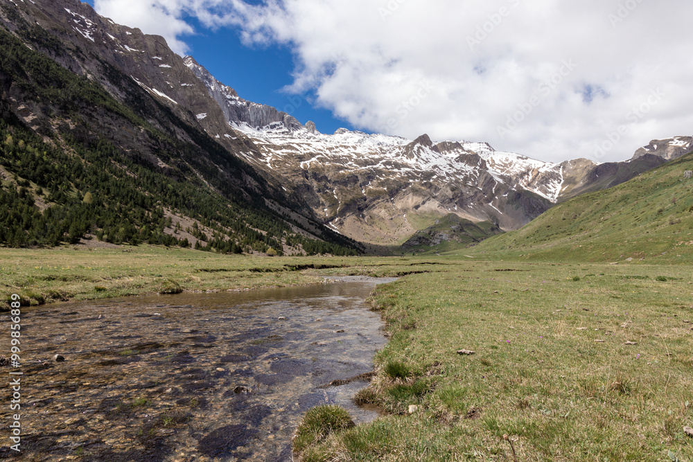 Expansive mountain valley with a clear stream flowing under snow-capped peaks and a partly cloudy sky Expansive mountain valley with a clear stream flowing under snow-capped peaks and a partly cloudy sky