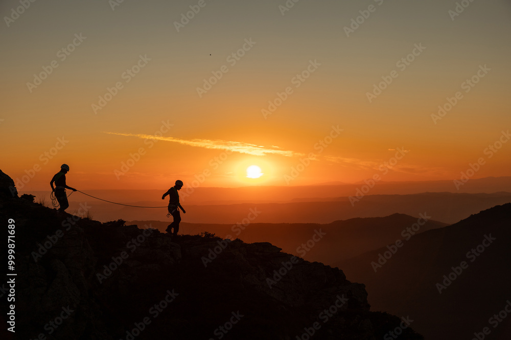 Two people are walking on a mountain side, with the sun setting in the background. Scene is peaceful and serene, as the two people enjoy the beautiful view of the sunset Two people are walking on a mountain side, with the sun setting in the background. Scene is peaceful and serene, as the two people enjoy the beautiful view of the sunset