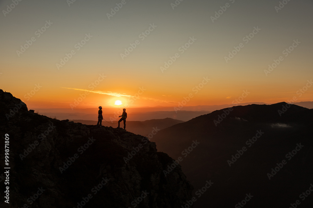 Two people are standing on a mountain top, looking out at the sunset. The sky is a mix of orange and pink hues, creating a serene and peaceful atmosphere Two people are standing on a mountain top, looking out at the sunset. The sky is a mix of orange and pink hues, creating a serene and peaceful atmosphere