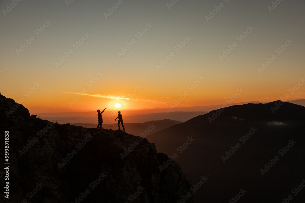 Two people are standing on a mountain top, looking at the sun as it sets. The sky is orange and the sun is low on the horizon Two people are standing on a mountain top, looking at the sun as it sets. The sky is orange and the sun is low on the horizon