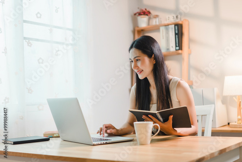 Young asian woman sits at a desk in a bright room working on her laptop while holding notebook. She enjoys a cup of coffee, radiating positivity.