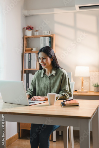 Young asian woman happily working on laptop in modern home office. The space is well-lit and features stylish decor, creating a productive atmosphere.