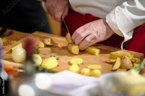 Obraz na płótnie Chef Slicing Potatoes on a Wooden Cutting Board During Culinary Preparation in a Warm, Ambient Light Kitchen Setting w salonie
