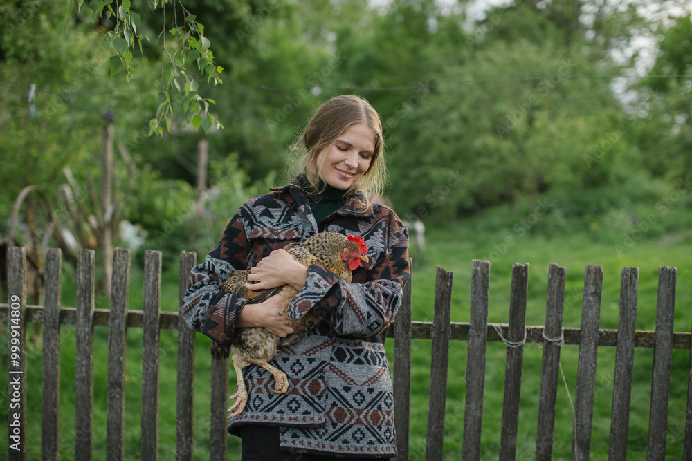 A young smiling woman holds a chicken in her hands while standing in a farm yard in the countryside. A young smiling woman holds a chicken in her hands while standing in a farm yard in the countryside.