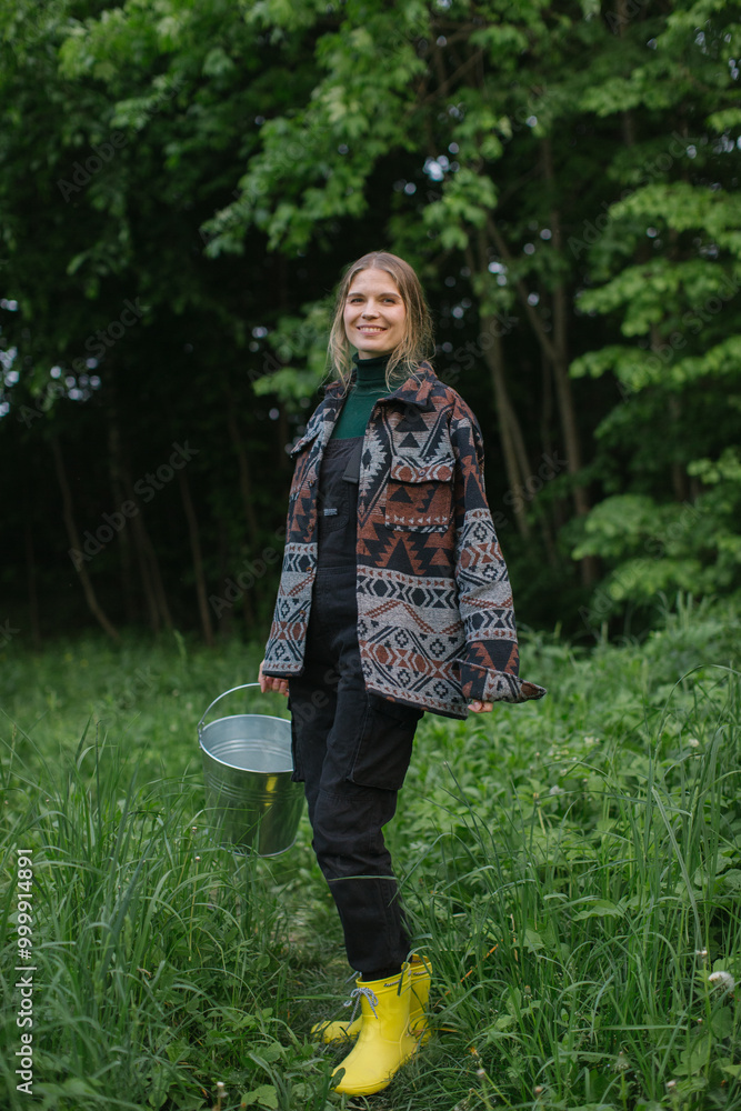 A young female farmer with a bucket in her hands poses against the background of deciduous trees in the village. A young female farmer with a bucket in her hands poses against the background of deciduous trees in the village.