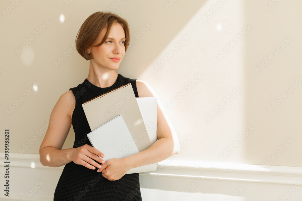 Young woman in a smart black dress standing by a wall holding blank folders and documents Young woman in a smart black dress standing by a wall holding blank folders and documents