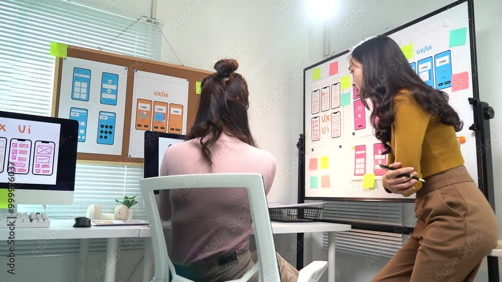 Two asian ui/ux web designers are discussing the design of a mobile application interface, using a whiteboard and sticky notes in their office Two asian ui/ux web designers are discussing the design of a mobile application interface, using a whiteboard and sticky notes in their office