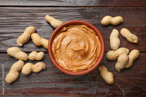 Tasty peanut butter in bowl and nuts on wooden table, flat lay