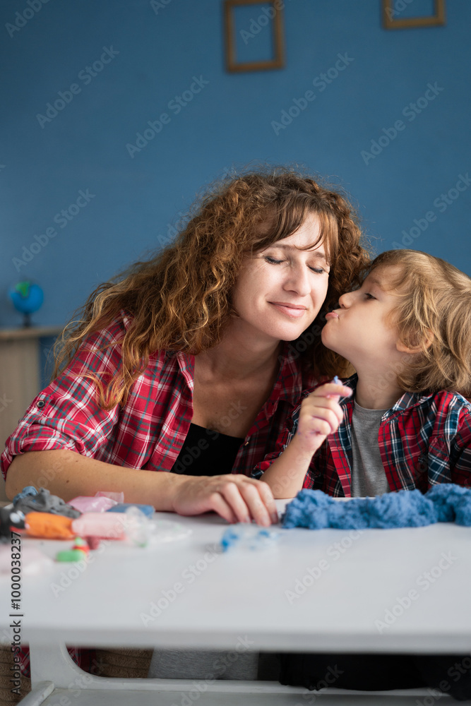 Cute little son kissing his mother and playing with kinetic sand sitting at table in the living room Cute little son kissing his mother and playing with kinetic sand sitting at table in the living room