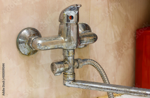 Old, corroded bathroom faucet with limescale buildup on beige tiled wall, showing neglect and poor maintenance.