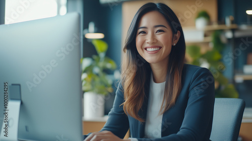 A happy Blasian female businesswoman sits at her desktop computer in a bright, creative office space. She smiles warmly during a video call meeting with a client, exuding confidenc photo