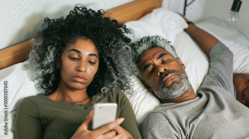  Afro-American couple lying in bed, the woman focused on her phone while the man is sleeping next to her. Emotional distance in the relationship