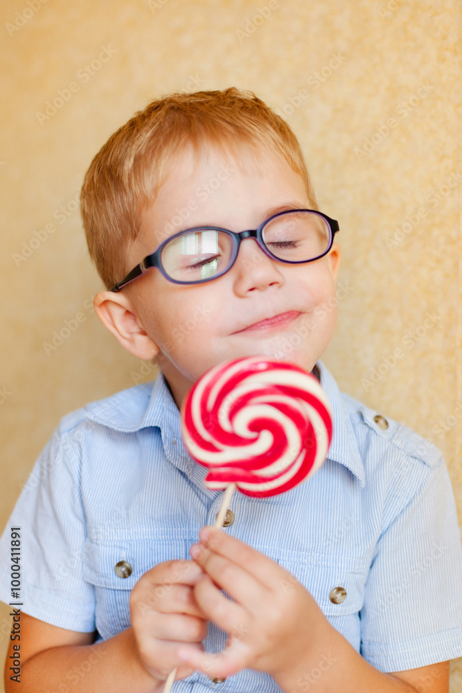 A young boy with glasses and squinting eyes is delightfully munching on a big red lollipop. A young boy with glasses and squinting eyes is delightfully munching on a big red lollipop.