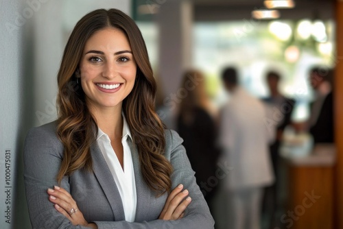 Wallpaper Mural Confident businesswoman with long brown hair stands in office corner, arms crossed and smiling at camera, wearing grey blazer and white shirt, with blurred colleagues in background. Torontodigital.ca