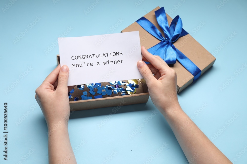 Woman holding card with phrase Congratulations You`re Winner and gift box on light blue background, closeup Woman holding card with phrase Congratulations You`re Winner and gift box on light blue background, closeup