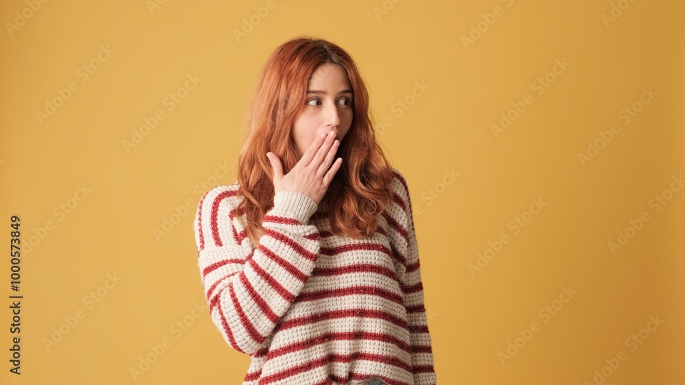 Surprised red-haired woman dressed in sweater looking at camera with big eyes, covering her mouth with her palm and looking to side on yellow background Surprised red-haired woman dressed in sweater looking at camera with big eyes, covering her mouth with her palm and looking to side on yellow background
