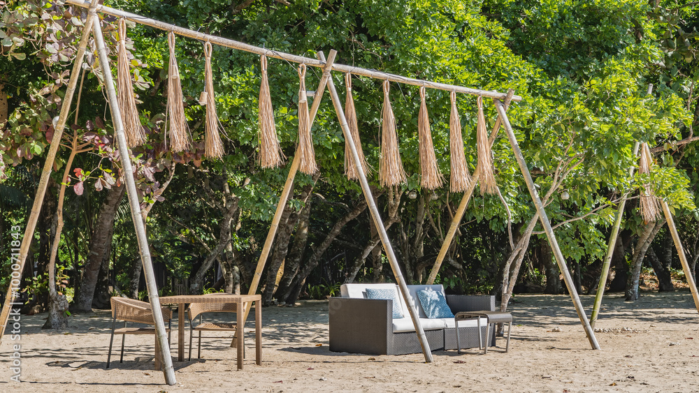 Fototapeta premium A relaxation area on a tropical beach. Wicker plastic furniture: tables, chairs, a sofa with soft cushions stand on the sand. Decorative tassels are suspended on bamboo poles. Lush green vegetation.