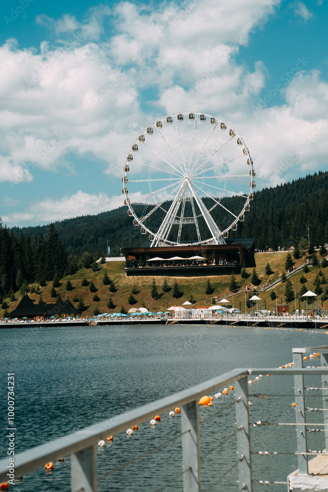 A Ferris wheel stands tall over a serene lake surrounded by lush trees on a clear, sunny day A Ferris wheel stands tall over a serene lake surrounded by lush trees on a clear, sunny day