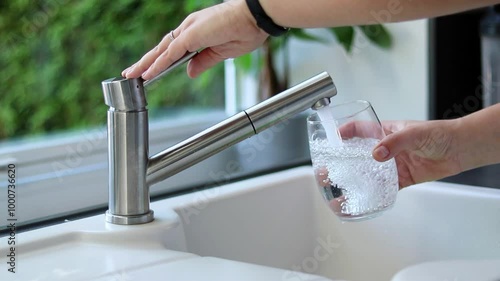 Close up of female hands pouring tap water into a glass in the kitchen. White sink and blurred background 