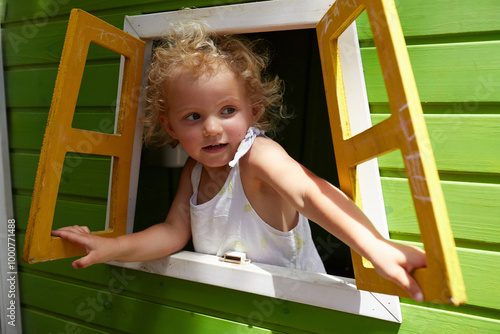 A young girl leans out of a playground cubby house window at something that caught her eye to the right
