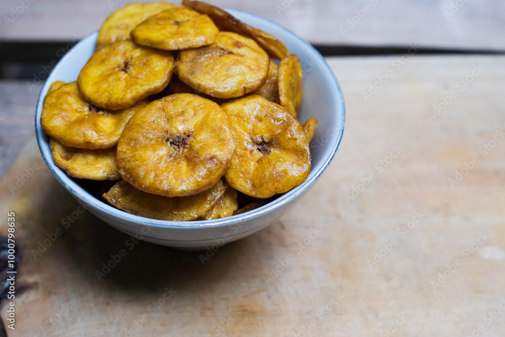 Keripik pisang or banana crackers on a white bowl at wooden cutting board. With copy space. Indonesian traditional snack made from banana and sugar. Keripik pisang or banana crackers on a white bowl at wooden cutting board. With copy space. Indonesian traditional snack made from banana and sugar.