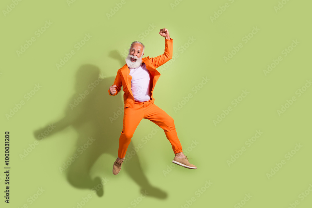Full length photo of funny showman jump raise fists wear orange suit isolated on green color background Full length photo of funny showman jump raise fists wear orange suit isolated on green color background