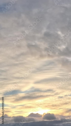 A cloudy sky at sunset. The clouds are predominantly white and gray with a subtle yellow hue. The setting sun is partially obscured by the clouds. photo