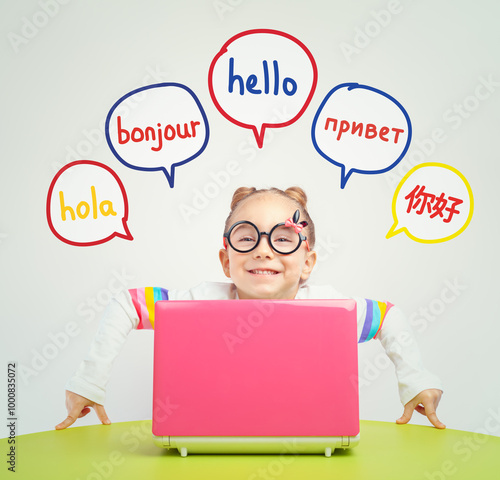 Foreign language learning concept of a beautiful cute little girl with eyeglasses behind a laptop on table and looking at camera. Hello word in Spanish, French, Russian and Chinese. 