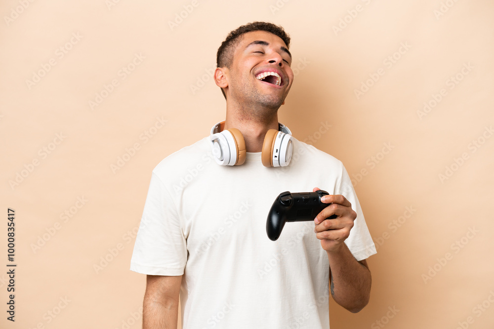 Young Brazilian man playing with a video game controller isolated on beige background laughing Young Brazilian man playing with a video game controller isolated on beige background laughing