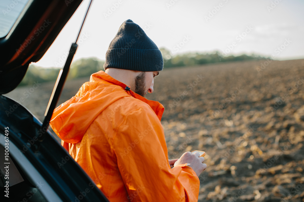 A male treasure hunter is standing near a car in a field with a phone in his hands. A male treasure hunter is standing near a car in a field with a phone in his hands.