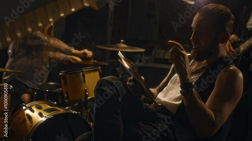 Medium shot of Caucasian male musician sitting on chair in recording studio nervously writing in notebook in company of bandmates adjusting percussion instruments and preparing for rehearsal photo