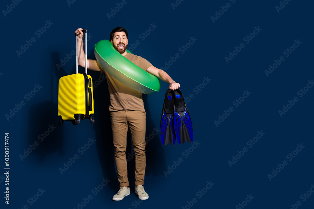 Photo of young funny man wear inflatable ring yellow suitcase and swimming flippers vacation preparation isolated on dark blue color background Photo of young funny man wear inflatable ring yellow suitcase and swimming flippers vacation preparation isolated on dark blue color background