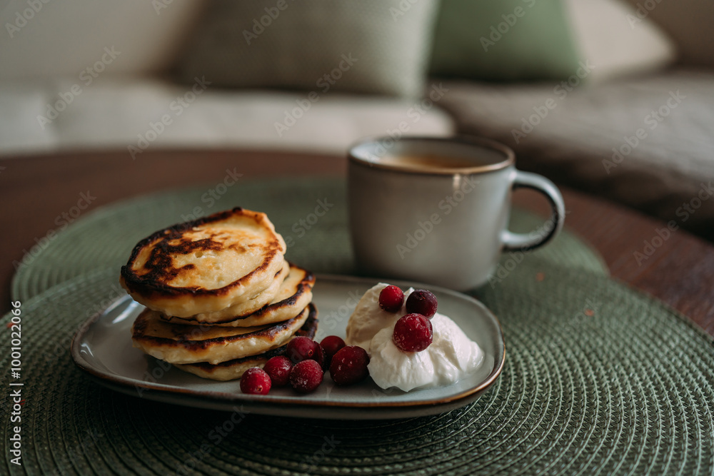 plate of homemade syrniki with whipped cream and cranberries, served with a cup of coffee on green woven placemat in cozy breakfast setting plate of homemade syrniki with whipped cream and cranberries, served with a cup of coffee on green woven placemat in cozy breakfast setting