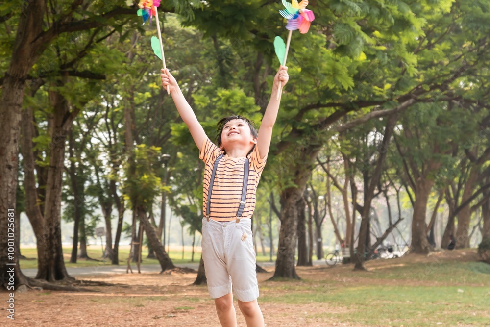Obraz premium Young Asian boy holding two colorful pinwheels in both hands, smiling happily outdoors in a park setting, wearing striped shirt with suspenders, sunny day surrounded by green trees, joyful expression