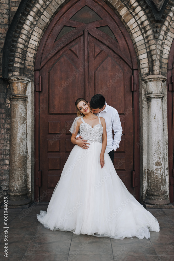 Stylish young groom and beautiful smiling bride curly blonde in white lace dress hugging, walking against the background of old brick church. Wedding photo, portrait of happy newlyweds. Stylish young groom and beautiful smiling bride curly blonde in white lace dress hugging, walking against the background of old brick church. Wedding photo, portrait of happy newlyweds.