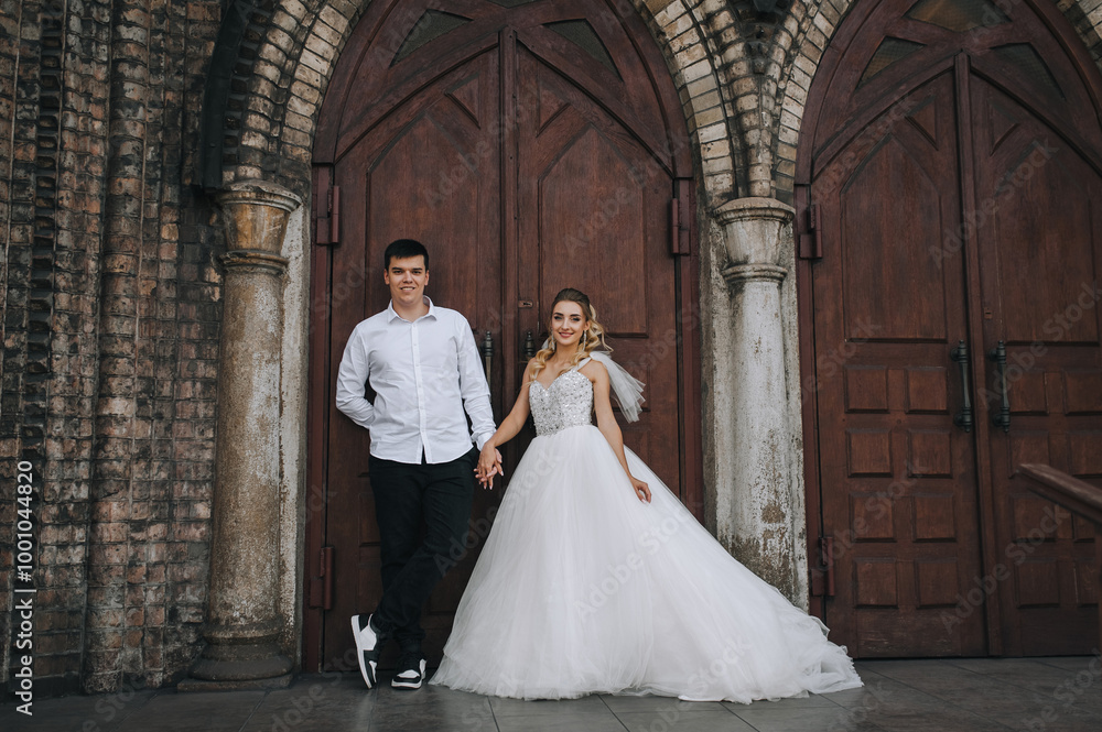 Fototapeta premium Stylish groom and beautiful bride curly blonde in white lace dress hugging, walking against the background of old brick church. Wedding photo, portrait of happy newlyweds.