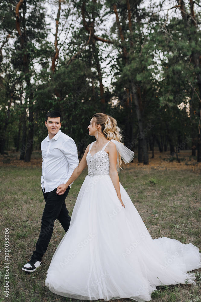 Stylish groom and beautiful smiling bride blonde with curly hair in white long dress walk together holding hands outdoors in pine forest in nature. Wedding photography, portrait of newlyweds. Stylish groom and beautiful smiling bride blonde with curly hair in white long dress walk together holding hands outdoors in pine forest in nature. Wedding photography, portrait of newlyweds.