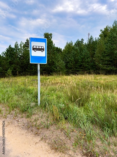 A bus stop sign stands on the side of a quiet rural road, surrounded by grassy fields and a backdrop of dense pine trees. The blue sky with scattered clouds adds a peaceful atmosphere to the scene