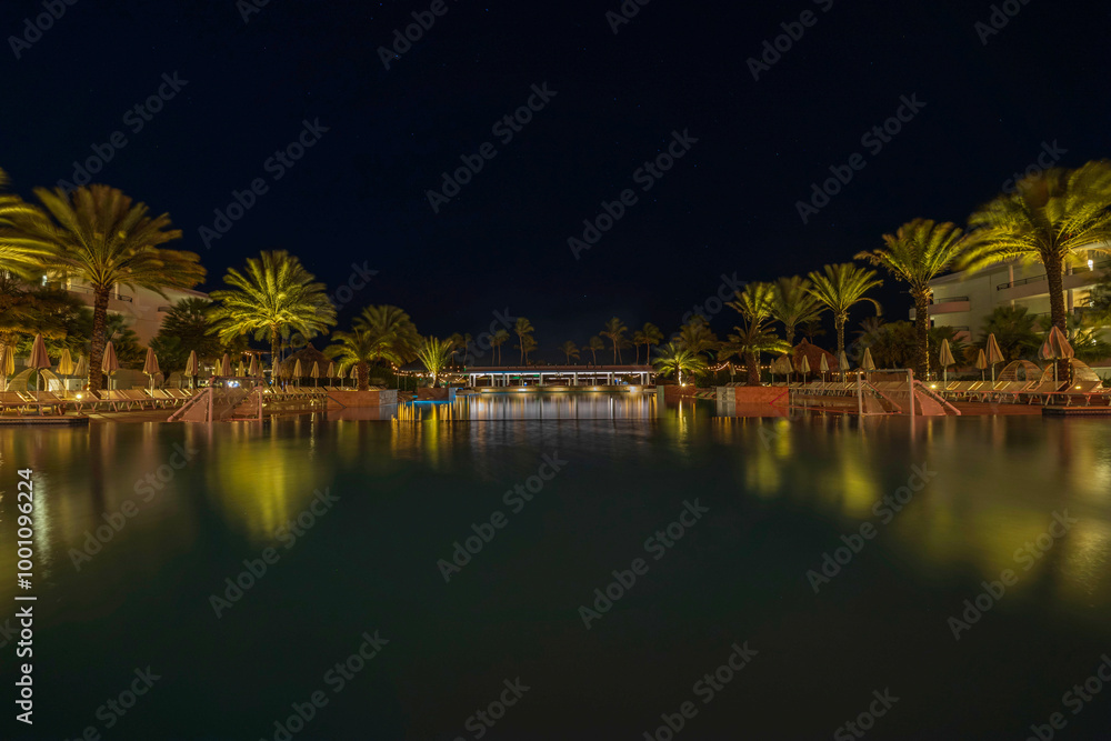 Night view of illuminated poolside with palm trees and lounge chairs at tropical resort in Curacao. Night view of illuminated poolside with palm trees and lounge chairs at tropical resort in Curacao.