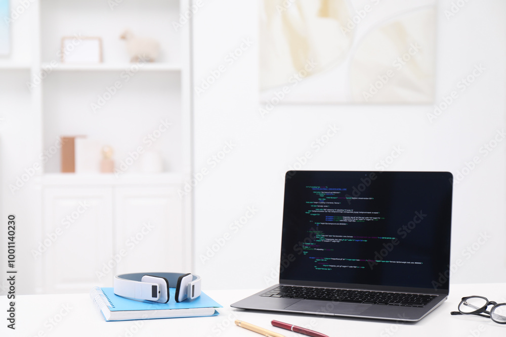 Fototapeta premium Programmer's workplace. Modern laptop with unfinished code, headphones and other supplies on white desk indoors