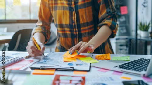 Person writing notes on a desk filled with sticky notes, laptops, and documents, depicting a busy and organized planning session in a brightly lit workspace.