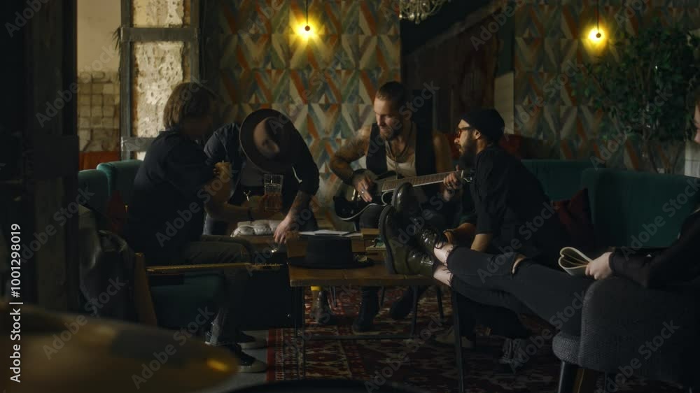 Full shot of diverse music group members greeting male guitarist wearing hat while sitting in bar at weekend, relaxing and having drinks before concer Full shot of diverse music group members greeting male guitarist wearing hat while sitting in bar at weekend, relaxing and having drinks before concer