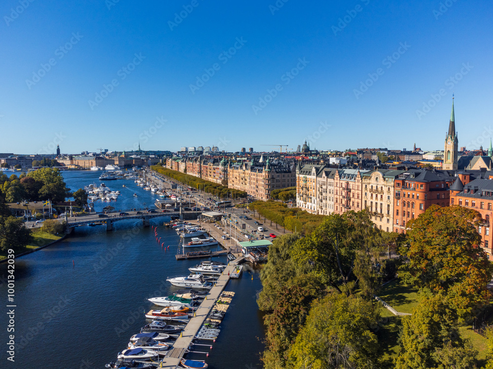 Fototapeta premium Strandvägen, famous street of Stockholm, early autumn, from above, with late 19th century buildings and an alley of green trees. Boats at dock. People walking